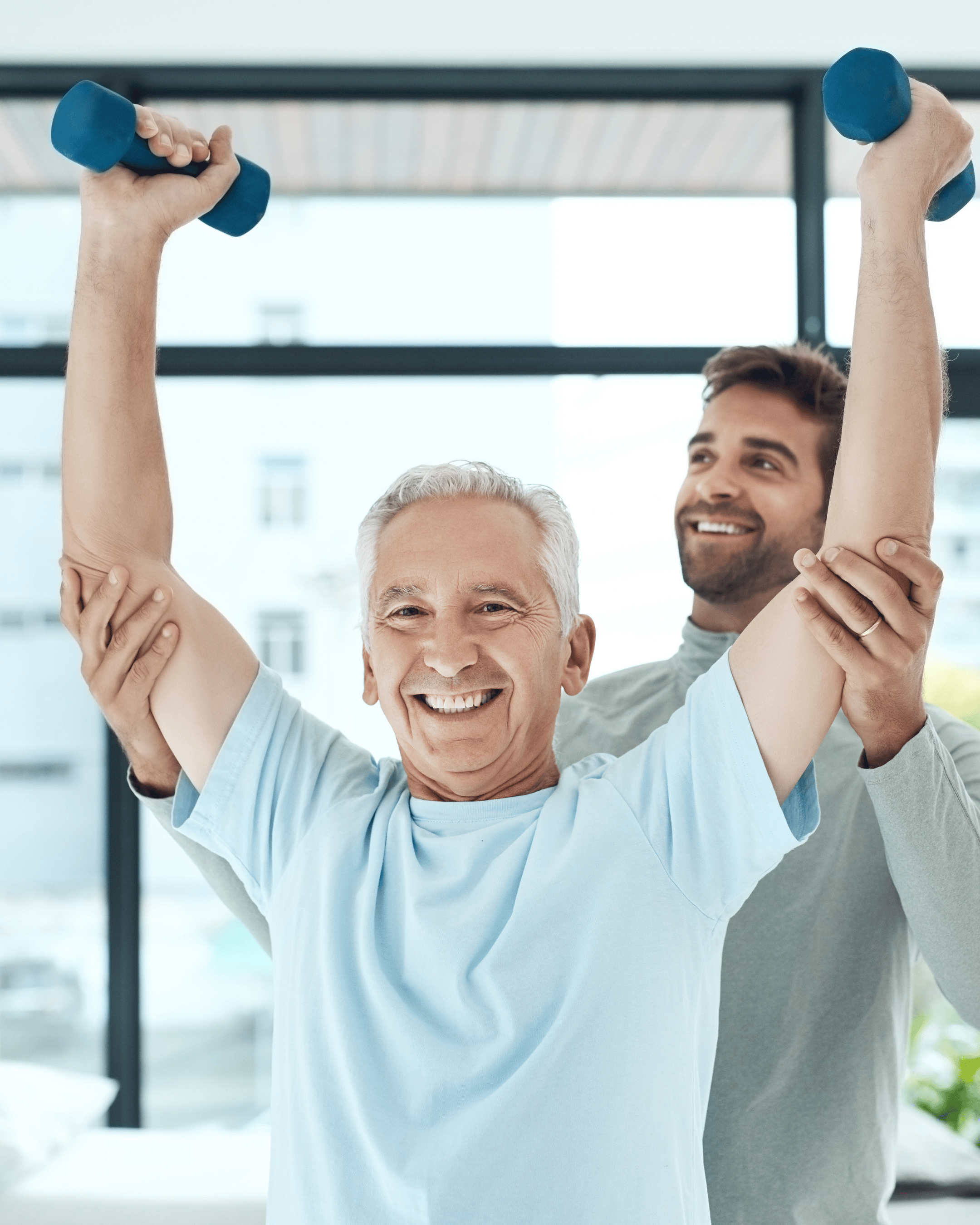 Senior man lifting dumbbells overhead with assistance from a physical therapist, both smiling during therapy session. Senior man lifting dumbbells overhead with assistance from a physical therapist, both smiling during therapy session.